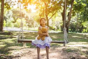 schattig klein meisje zit schommels, teddybeer zit met haar, klein meisje wees naar de boom. foto