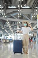 jonge vrouw met gezichtsmasker met bagage die op de luchthaven loopt, bescherming tegen infectie met coronavirusziekte, aziatische vrouwelijke reiziger met hoed. tijd om te reizen na het concept van de boosterdosis van het vaccin foto
