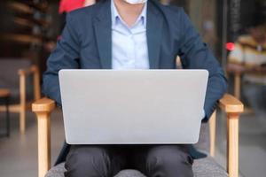 jonge zakenman in pak met chirurgisch gezichtsmasker en met behulp van laptop, man toetsenbord computer notebook typen in kantoor of café. covid-19 pandemie, technologie en nieuw normaal concept foto