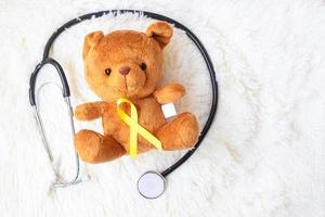 geel lint op beer pop met stethoscoop op witte achtergrond voor het ondersteunen van kind leven en ziekte. september kinderkanker bewustzijn maand en wereld kanker dag concept foto