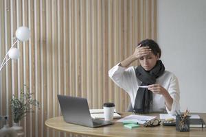 vermoeide dame freelancer zit aan bureau op kantoor aan huis ziekelijk kijkend naar digitale thermometer foto