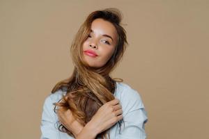 portret van doordachte mooie vrouw kijkt opzij, denkt aan iets aangenaams, steekt haar over nek, draagt make-up, stijlvol shirt, geïsoleerd op bruine achtergrond. mensen, schoonheid, levensstijl foto