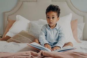 schattige Afro-Amerikaanse kleine jongen met krullend haar zittend op bed met boek foto