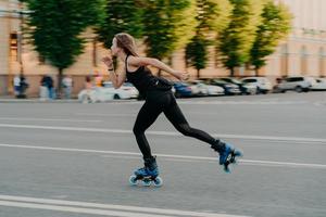 professionele vrouwelijke roller demonstreert haar capaciteiten van skaten rijdt heel snel op de weg langs de stad geniet van een zonnige dag gekleed in zwarte actieve kleding. actieve levensstijl hobby en fitness activiteit foto