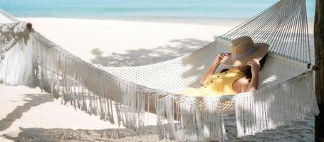 gelukkige vrouw reiziger is ontspannen in een hangmat op het paradijselijke strand. vrouwelijke toerist in gele jurk rust in de buurt van tropische zee. vakantie, reizen, zomer, reislust en vakantieconcept foto