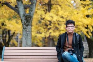 gelukkige man geniet in het park buiten in het herfstseizoen, aziatische reiziger in jas en camera tegen gele ginkgo-bladerenachtergrond foto