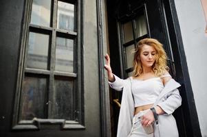 stijlvolle krullend blond model meisje slijtage op wit poseren tegen oude houten deur. foto