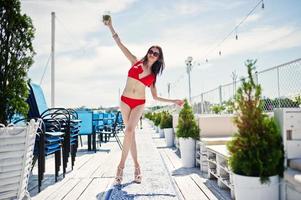 geweldige jonge vrouw in rode bikini poseren met een mojito-cocktail in haar hand op de kade bij het meer. foto