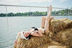 portret van een prachtig meisje in zwarte bikini zwembroek poseren op de hooibaal met een hoed aan het meer. foto