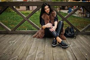 fashion outdoor foto van prachtige sensuele vrouw met donker haar in elegante kleding en luxe bontjas zittend tegen houten balustrades.