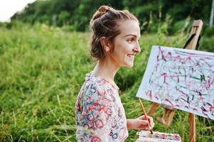 portret van een prachtige gelukkige jonge vrouw in een mooie jurk zittend op het gras en schilderen op papier met aquarellen. foto