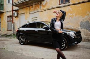 elegant blond meisje draagt een zwarte leren jas die zich voordeed in de straten van de stad achtergrond luxeauto. foto