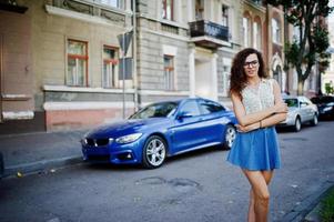 krullend stijlvol meisje draagt een blauwe jeansrok, blouse en bril in de buurt van blauwe auto op straat van de stad. foto