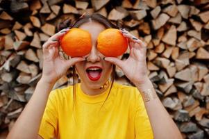 jong grappig meisje met lichte make-up, draag op geel shirt, houd twee oranje in de ogen tegen houten achtergrond. foto