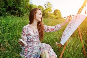 portret van een prachtige gelukkige jonge vrouw in een mooie jurk zittend op het gras en schilderen op papier met aquarellen. foto