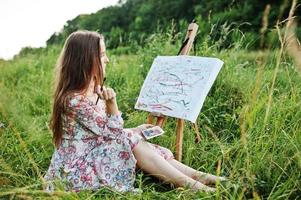 portret van een prachtige gelukkige jonge vrouw in een mooie jurk zittend op het gras en schilderen op papier met aquarellen. foto