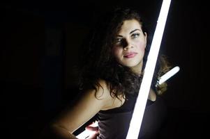 krullend brunette plus size model met twee lange led-lampen op zwarte kamer. foto