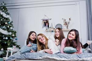vier schattige vriendenmeisjes dragen warme truien en zwarte broeken op bed op nieuwjaarskamer op studio, spelen met kussens. foto