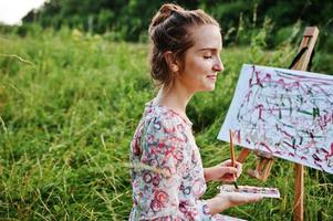 portret van een prachtige gelukkige jonge vrouw in een mooie jurk zittend op het gras en schilderen op papier met aquarellen. foto