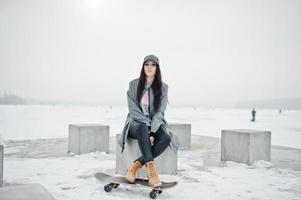 stijlvolle brunette meisje in grijze pet, casual straatstijl met skate board op winterdag. foto