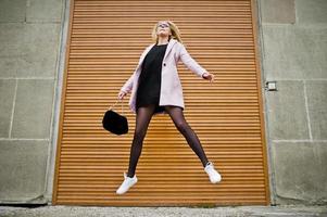 blond meisje met bril en roze jas met handtas springen tegen luiken. foto