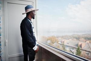 rijke afrikaanse man kijkt naar panoramisch raam in zijn penthouse. portret van succesvolle zwarte man in hoed binnen. foto