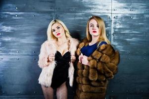 twee elegante blonde meisjes dragen een bontjas en een combi-jurk tegen een stalen muur in de studio. foto