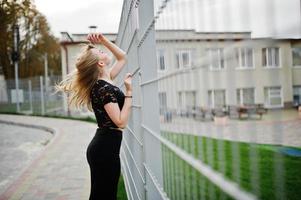 elegant blond meisje draagt zwarte poseren in straten van de stad tegen ijzeren kooi. foto
