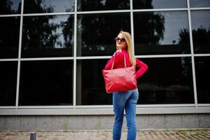 portret van een fantastische jonge vrouw in rode blouse en spijkerbroek poseren met haar handtas en zonnebril buiten het winkelcentrum op glazen achtergrond. foto