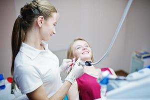 aantrekkelijke patiënt in rood-violette jurk die op de tandartsstoel ligt terwijl vrouwelijke tandarts haar tanden behandelt met speciale instrumenten.
