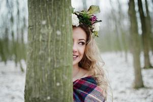 krullend schattig blond meisje met krans in geruite plaid op besneeuwde bos in winterdag. foto