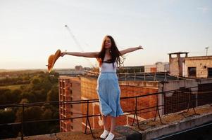 portret van een mooi meisje in casual kleding poseren op het dak met haar hoed in haar hand bij de zonsondergang. foto