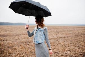 portret van brunette krullend meisje in jeans jasje met zwarte paraplu op veld. foto