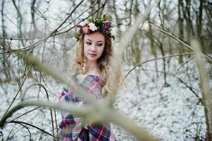 krullend schattig blond meisje met krans in geruite plaid op besneeuwde bos in winterdag. foto