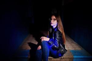 nachtportret van meisjesmodel op bril, jeans en leren jas, met blauwe slinger op haar. foto