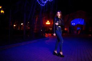 nachtportret van meisjesmodel op bril, jeans en leren jas tegen blauwe lichtenslinger van stadsstraat. foto