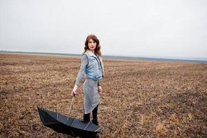 portret van brunette krullend meisje in jeans jasje met zwarte paraplu op veld. foto