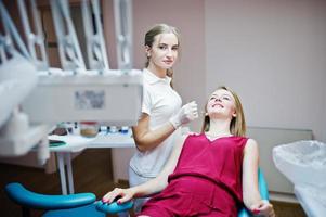 aantrekkelijke patiënt in rood-violette jurk die op de tandartsstoel ligt terwijl vrouwelijke tandarts haar tanden behandelt met speciale instrumenten.