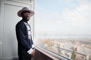 rijke afrikaanse man kijkt naar panoramisch raam in zijn penthouse. portret van succesvolle zwarte man in hoed binnen. foto