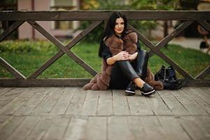 fashion outdoor foto van prachtige sensuele vrouw met donker haar in elegante kleding en luxe bontjas zittend tegen houten balustrades.