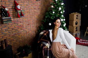 brunette meisje in jurk gesteld in de buurt van nieuwjaarsboom met kerstversiering in baksteen studio kamer. foto