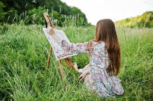 portret van een prachtige gelukkige jonge vrouw in een mooie jurk zittend op het gras en schilderen op papier met aquarellen. foto