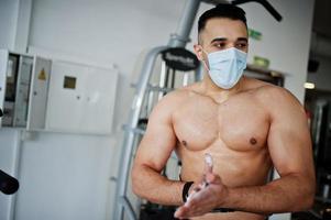 gespierde arabische man, draag een medische gezichtsmaskertraining in een moderne sportschool. fitness arabische mannen met naakte torso die oefeningen doen, tijdens een pandemie van het coronavirus. foto