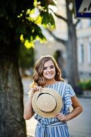 portret van een zeer aantrekkelijke jonge vrouw in gestreepte overall poseren met haar hoed op een stoep in een stad met bomen op de achtergrond. foto