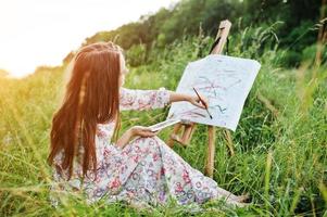 portret van een prachtige gelukkige jonge vrouw in een mooie jurk zittend op het gras en schilderen op papier met aquarellen. foto