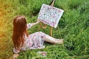 portret van een prachtige gelukkige jonge vrouw in een mooie jurk zittend op het gras en schilderen op papier met aquarellen. foto