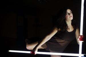 krullend brunette plus size model met twee lange led-lampen op zwarte kamer. foto