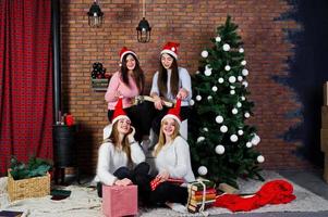 vier schattige vriendenmeisjes dragen warme truien, zwarte broeken en kerstmutsen tegen nieuwjaarsboom met kerstversiering in de studio. foto