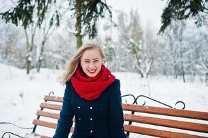 blond meisje in rode sjaal en jas zittend op een bankje op winterdag. foto