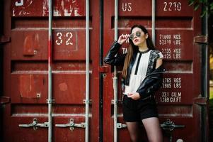 stijlvolle brunette meisjes dragen op leren jas en korte broek, zonnebril tegen zeecontainer. foto